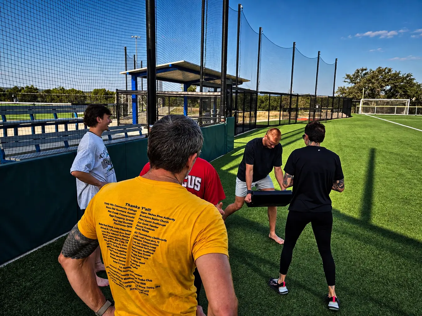 A small group training outdoors on a turf field, one member bracing a strike pad while the others work through the drill